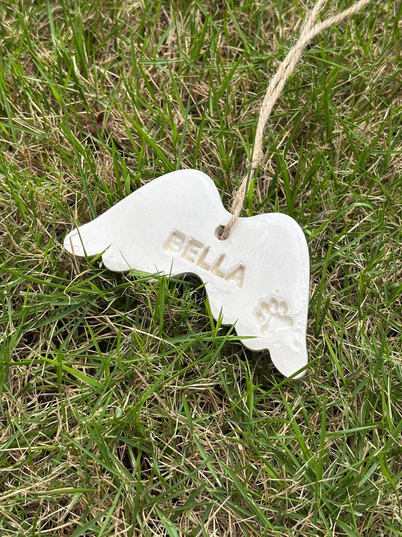 K&ouml;nnte beinhalten: Ein wei&szlig;er Keramik-Engelsfl&uuml;gel-Ornament mit dem Namen "BELLA" und einem Pfotenabdruck. Das Ornament h&auml;ngt an einer Schnur aus Naturfaser vor dem Hintergrund von gr&uuml;nem Gras.
