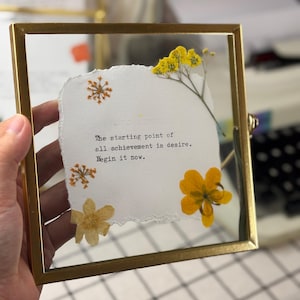 May include: A framed artwork with a gold-colored metal frame. The artwork features a piece of torn white paper with the quote "The starting point of all achievement is desire. Begin it now." Dried yellow flowers and orange accents are arranged around the paper.