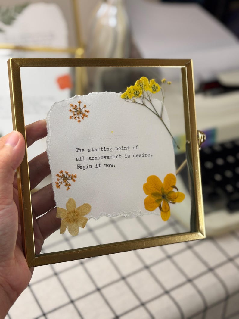 May include: A framed artwork with a gold-colored metal frame. The artwork features a piece of torn white paper with the quote "The starting point of all achievement is desire. Begin it now." Dried yellow flowers and orange accents are arranged around the paper.
