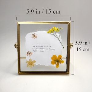 May include: A square, gold-toned framed piece featuring dried flowers and a quote. The frame is 5.9 inches (15 cm) square. The quote reads, "The starting point of all achievement is desire. Begin it now." Yellow and white flowers are arranged on a white paper.