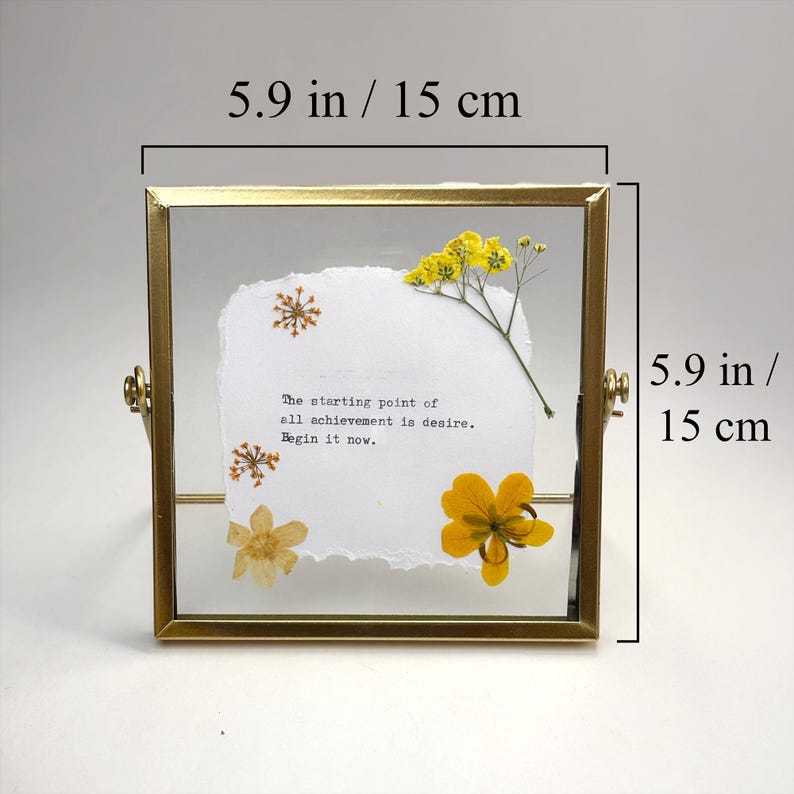 May include: A square, gold-toned framed piece featuring dried flowers and a quote. The frame is 5.9 inches (15 cm) square. The quote reads, "The starting point of all achievement is desire. Begin it now." Yellow and white flowers are arranged on a white paper.