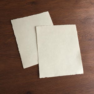 May include: Two rectangular sheets of off-white paper with uneven, deckled edges. The paper has a textured, fibrous surface and is arranged in an overlapping position, displaying a light cream colour throughout the entire surface area.