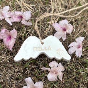K&ouml;nnte beinhalten: Wei&szlig;es Keramik-Engelsfl&uuml;gel-Ornament mit dem Namen "BELLA" und einem Pfotenabdruck. Das Ornament h&auml;ngt an einer Schnur aus Naturfaser und ist von rosa Bl&uuml;ten auf trockenem Gras umgeben.
