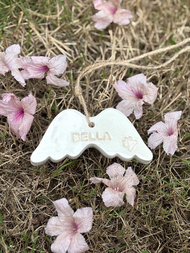 K&ouml;nnte beinhalten: Wei&szlig;es Keramik-Engelsfl&uuml;gel-Ornament mit dem Namen "BELLA" und einem Pfotenabdruck. Das Ornament h&auml;ngt an einer Schnur aus Naturfaser und ist von rosa Bl&uuml;ten auf trockenem Gras umgeben.