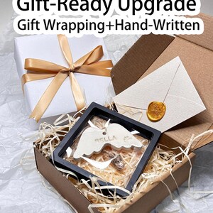 K&ouml;nnte beinhalten: Eine Geschenkbox mit wei&szlig;em Deckel und goldenem Band, eine braune Box mit einem Geschenk darin und ein wei&szlig;er Umschlag mit einem goldenen Siegel. Das Geschenk in der braunen Box ist ein wei&szlig;er Engelsfl&uuml;gel mit dem Namen "BELLA". Text auf dem Bild lautet "Gift-Ready Upgrade".
