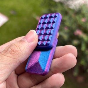 May include: A two-part fidget toy in shades of purple, blue, and pink. The top section has a textured surface with pyramid-shaped bumps. The bottom section features a geometric design with colour gradients. The toy is held in a hand.