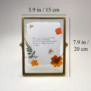 May include: A brass-framed photo holder, 5.9 inches wide and 7.9 inches tall, with a clear glass front. Inside, a white paper displays pressed flowers and text: "Home is not where you are from..." Decorative home decor.