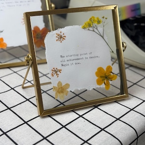May include: A square, gold-framed glass picture frame with a white paper insert. The paper has the text "The starting point of all achievement is desire. Begin it now." and is decorated with dried yellow flowers.