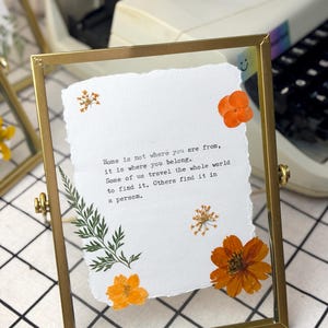May include: A rectangular gold-colored metal frame with a clear glass front, holding a piece of textured white paper. The paper has the quote "Home is not where you are from, it is where you belong." Orange dried flowers and green leaves are arranged around the paper.