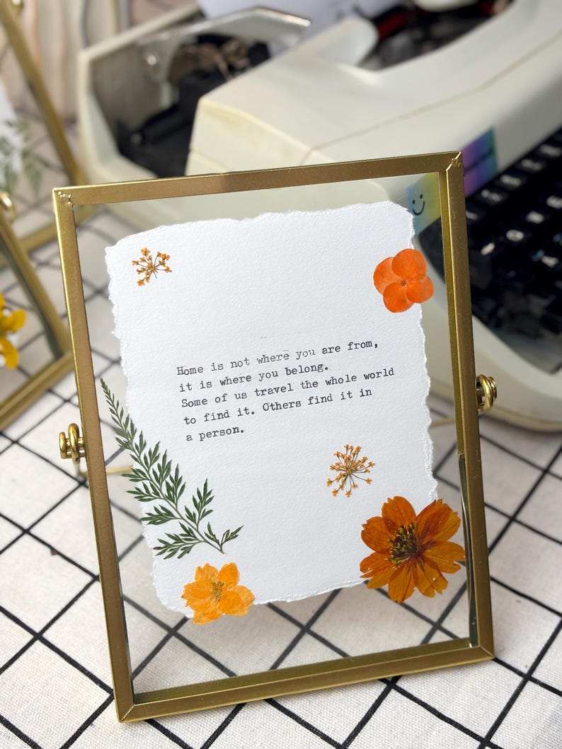 May include: A rectangular gold-colored metal frame with a clear glass front, holding a piece of textured white paper. The paper has the quote "Home is not where you are from, it is where you belong." Orange dried flowers and green leaves are arranged around the paper.