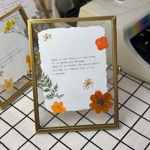 May include: A framed piece of art with a gold metal frame. The artwork features a white paper with the quote "Home is not where you are from, it is where you belong." Orange flowers and green leaves are arranged around the text.