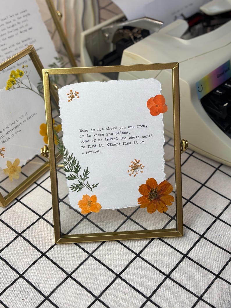 May include: A framed piece of art with a gold metal frame. The artwork features a white paper with the quote "Home is not where you are from, it is where you belong." Orange flowers and green leaves are arranged around the text.
