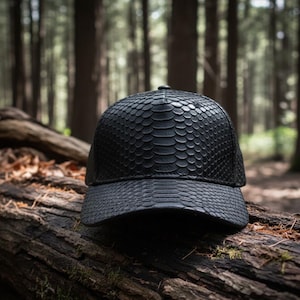 Puede incluir: Gorra de béisbol negra con un patrón texturizado de piel de serpiente. La gorra está sobre un tronco desgastado en un entorno forestal. La gorra tiene una visera curva y un botón en la parte superior.