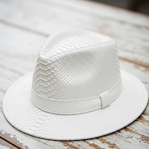 Può includere: Un cappello fedora bianco con una trama a pelle di serpente. Il cappello presenta una corona strutturata, un'ampia tesa e una fascia attorno alla base della corona. Il cappello è realizzato in un materiale strutturato.