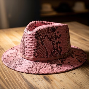 Può includere: Un cappello fedora rosa con un motivo a pelle di serpente. Il cappello presenta una fascia in pelle marrone intorno alla corona e una piccola fibbia. Il cappello è appoggiato su una superficie di legno.