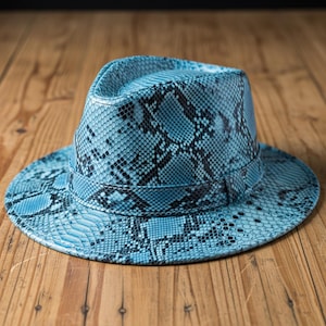 Può includere: Un cappello fedora con un motivo a pelle di serpente nei toni del blu e del nero. Il cappello ha una corona strutturata e un'ampia tesa. Il cappello è realizzato in un materiale strutturato e viene esposto su una superficie di legno.