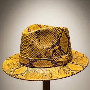Può includere: Un cappello fedora giallo con un motivo a pelle di serpente. Il cappello presenta un'ampia tesa e una fascia attorno alla corona. Il motivo a pelle di serpente è un mix di giallo e marrone, creando un aspetto strutturato. Il cappello è esposto su un supporto di legno.
