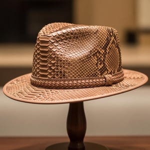 May include: A brown fedora hat with a snakeskin pattern. The hat features a wide brim and a braided leather band around the crown. The hat is displayed on a wooden stand.