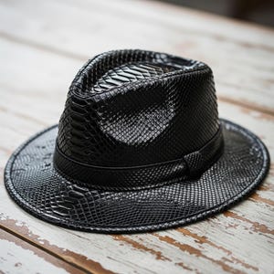 Może przedstawiać: Czarny kapelusz typu fedora o fakturze skóry węża. Kapelusz ma strukturalną koronę, szerokie rondo i opaskę wokół podstawy korony. Kapelusz wykonany jest z błyszczącego, teksturowanego materiału.