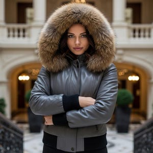 May include: A gray leather jacket with a fur-trimmed hood. The jacket has a bomber style with black ribbed cuffs and hem. The fur trim is a mix of brown and tan colors. The jacket is buttoned up and the person is wearing a black top.