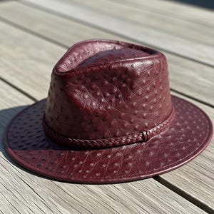 Puede incluir: Sombrero fedora de cuero burdeos con superficie texturizada y banda trenzada. El sombrero tiene ala ancha y forma de corona clásica. El sombrero está sobre una superficie de madera. Las medidas son en centímetros.