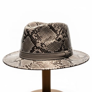 May include: A snakeskin-patterned fedora hat in grey and black. The hat has a grey band around the crown and a wide brim. It is displayed on a wooden stand against a white backdrop. The hat's design is reminiscent of reptile skin.