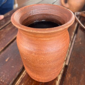 May include: A terracotta-colored ceramic mug filled with a dark liquid, possibly coffee or tea. The mug has a wide, slightly flared rim and a textured surface. It sits on a wooden table, with another similar mug visible in the background.