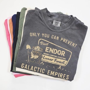 May include: A pile of t-shirts in various colours. The top shirt is dark grey and features the text "ONLY YOU CAN PREVENT Visit ENDOR Hiking-Camping-Feasting Lunar Forest GALACTIC EMPIRES" in tan. Other colours visible are pink, orange, navy, black, green, and white.