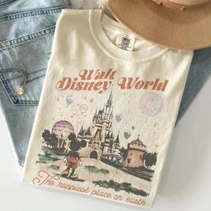 Może przedstawiać: Kremowa koszulka z grafiką w stylu vintage z Walt Disney World, z Myszka Miki, zamkiem i frazą "The happiest place on earth". Koszulka jest połączona z jasnoniebieskimi dżinsami i naszyjnikiem.