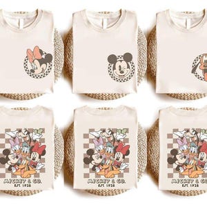 May include: Cream-coloured t-shirts featuring Disney characters. Designs include Minnie Mouse, Mickey Mouse, and Pluto in checkered circles, and a group of characters with "MICKEY & CO. EST 1928". The shirts are folded on woven mats.
