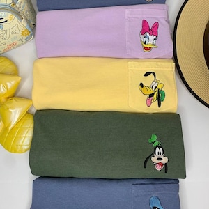 以下が含まれることがあります： ミッキーマウス、デイジーダック、プルート、グーフィー、ドナルドダックが刺繍されたポケットが付いた、5枚のカラフルな長袖シャツのスタック。 シャツは、青、ラベンダー、黄色、緑、ネイビーの色合いです。