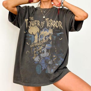 May include: Dark gray t-shirt featuring the text "The Tower of Terror" above an illustration of a building and Mickey Mouse in a bellhop uniform. The design includes bats and the words "Hollywood Tower".