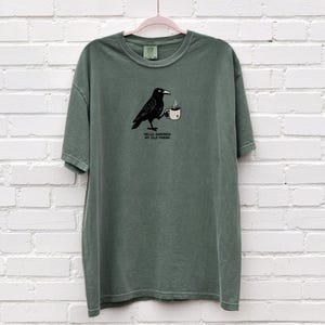 Peut inclure: T-shirt vert sauge avec un dessin d'un corbeau noir tenant une tasse à café. Le texte "Hello Darkness My Old Friend" est imprimé en dessous. Le t-shirt est suspendu à un cintre rose devant un mur de briques blanches.