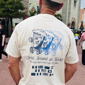 Puede incluir: Camiseta color crema con un diseño azul y blanco de jarras de cerveza de dibujos animados. El texto en la camiseta dice "Drink Around the World" y "From First Sip to Last Call in 1.2 Miles". Debajo del texto hay varias banderas de países.