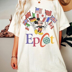 Puede incluir: Camiseta color crema con Mickey Mouse sosteniendo banderas de varios países. La palabra "Epcot" está impresa en letras coloridas debajo del gráfico. La camiseta tiene mangas cortas y un corte holgado.