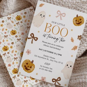 May include: Two Halloween-themed invitation cards with a pumpkin and ghost design. The main card reads "Our Little Boo is turning two" with event details. The cards are set on a woven basket and a cream-colored knitted blanket.