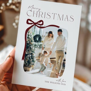 Könnte beinhalten: Eine weiße Weihnachtskarte mit dem Schriftzug "Merry Christmas" in einer Schreibschrift, umrahmt von einem bordeauxfarbenen Schleifendesign. Die Karte zeigt ein Familienfoto innerhalb des Bandes, mit dem Text "with love" und einem Namen und Jahr unten.