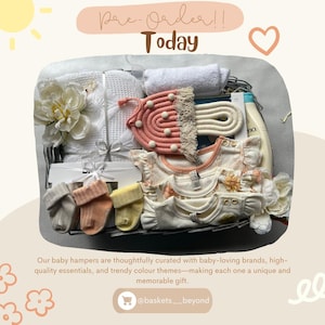 Op de afbeelding: Een babycadeaumand gevuld met babykleding, sokken, een washandje en lotion. De mand bevat een witte deken, een roze en crème macrame-item en bloemen. Tekst luidt "Pre-Order!! Today".