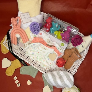 May include: A white wicker basket filled with baby items, including a yellow blanket, a peach-colored rainbow, baby clothes, socks, and bath toys. The basket also contains wooden heart cutouts and a bottle of baby wash. The background is red.