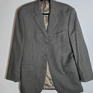 May include: A grey blazer featuring a three-button front closure, notched lapels, and two flap pockets at the waist. The garment includes a single chest pocket and is lined with a smooth, light brown fabric visible at the interior collar and front.