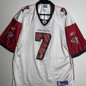 May include: A white Reebok Atlanta Falcons football jersey featuring the number 7 in red with black trim on the front and shoulders. The sleeves are red with black cuffs and white Reebok logos. The word FALCONS is printed above the number 7.