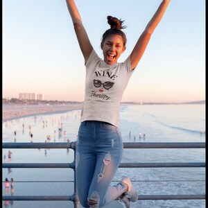 Pu&ograve; includere: Una persona salta in aria, indossa una maglietta bianca con le parole "WINE Therapy" e una grafica di occhiali da sole. La persona indossa jeans blu con ginocchia strappate e scarpe da ginnastica grigie. Lo sfondo mostra un molo e l'oceano.