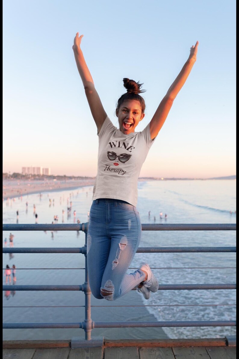 Pu&ograve; includere: Una persona salta in aria, indossa una maglietta bianca con le parole "WINE Therapy" e una grafica di occhiali da sole. La persona indossa jeans blu con ginocchia strappate e scarpe da ginnastica grigie. Lo sfondo mostra un molo e l'oceano.