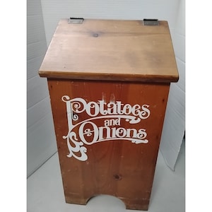 May include: A wooden potato and onion storage bin with a hinged lid. The bin is stained a warm brown color and features the words "Potatoes and Onions" in white script on the front. The bin has a rectangular shape with a slightly angled top.