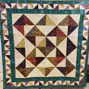 May include: A handmade quilt with a geometric triangle pattern in cream, burgundy, gold, and purple. It has a teal border and a cream background. The quilt's design creates a cozy and colourful aesthetic, suitable for a bed or sofa.