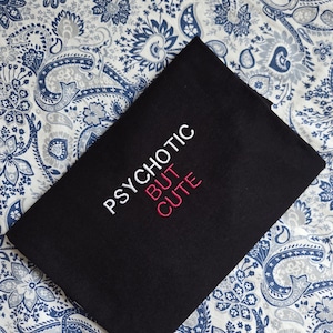 Może przedstawiać: Czarna koszulka z wyhaftowanym napisem PSYCHOTIC białą nicią oraz słowem BUT CUTE różową nicią. Odzież jest złożona na płasko, eksponując wyraziste, wyśrodkowane litery na tle jednolitego czarnego materiału koszulki.