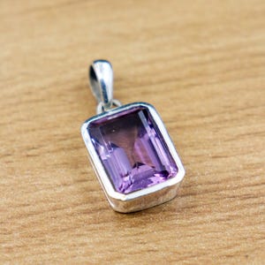 Pode incluir: Um pingente retangular de ametista com moldura prateada. A pedra preciosa é de cor roxa escura e é colocada em uma moldura prateada polida. O pingente tem uma pequena argola na parte superior para uma corrente.