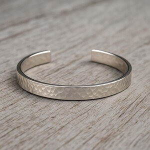 Könnte beinhalten: Ein silbernes Armreif-Armband mit gehammerter Textur. Das offene Armband ist etwa 0,6 cm breit und liegt auf einer Holzoberfläche. Das Armband hat ein schlichtes, modernes Design.