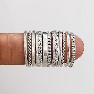 Może przedstawiać: Kolekcja srebrnych pierścionków o różnych wzorach, w tym skręcone, wzorzyste i gładkie obrączki. Pierścionki są prezentowane na palcu, prezentując różne tekstury i style. Pierścionki wykonane są z błyszczącego metalu.