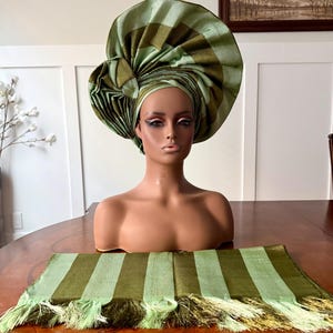 May include: A mannequin head wearing a large, intricately folded headwrap in alternating shades of olive green and light sage green. A matching rectangular fabric piece with fringed edges in the same striped green pattern lies in the foreground.